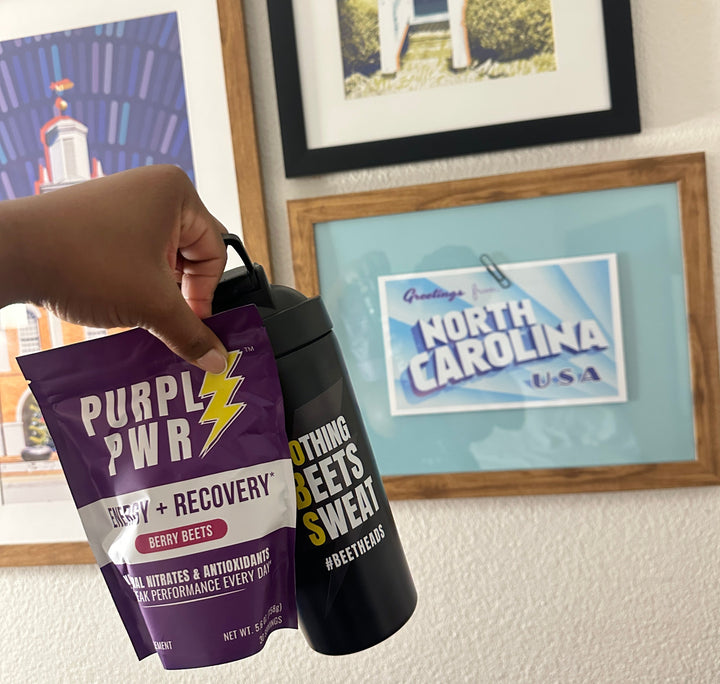 Hand holding a Purpl PWR supplement package with framed pictures in the background
